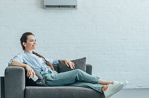 happy woman relaxing on sofa with air conditioner on white wall summer heat concept - Trusted Los Angeles HVAC Service Econo Air HVAC ventilation and air conditioning heating systems